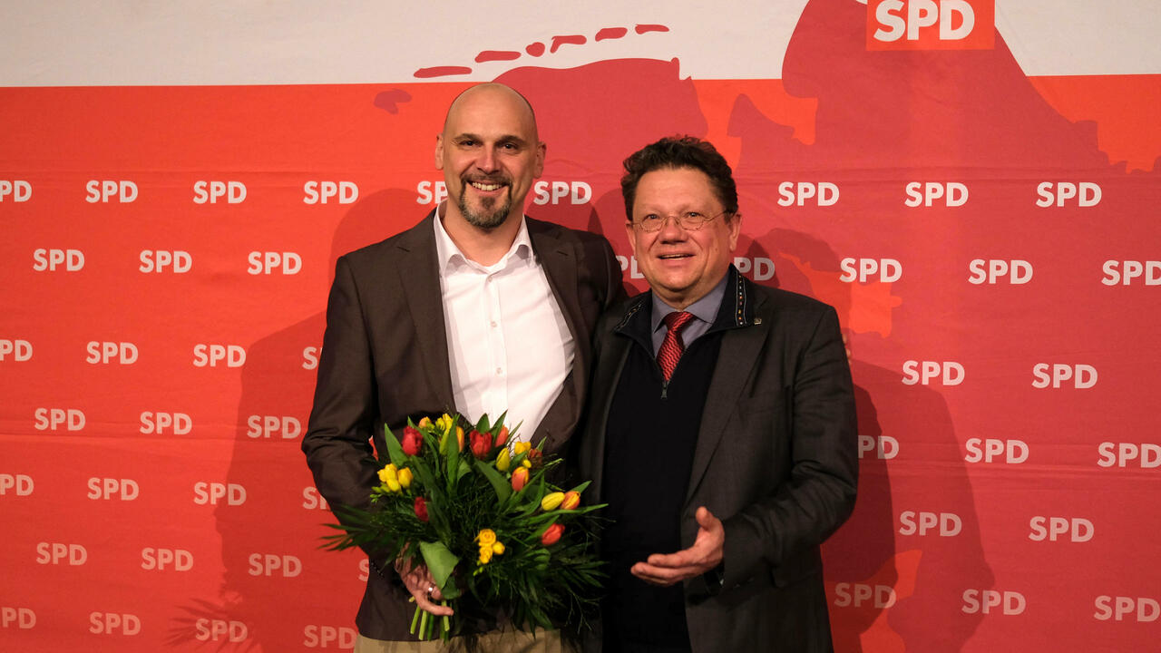 Dr. Andreas Philippi gratuliert Alexander Saade mit einem Blumenstrauß vor einem SPD Plakat.