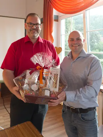 Zwei Männer lächeln in die Kamera und halten gemeinsam einen großen, dekorativ verpackten Geschenkkorb bei einer Übergabe.