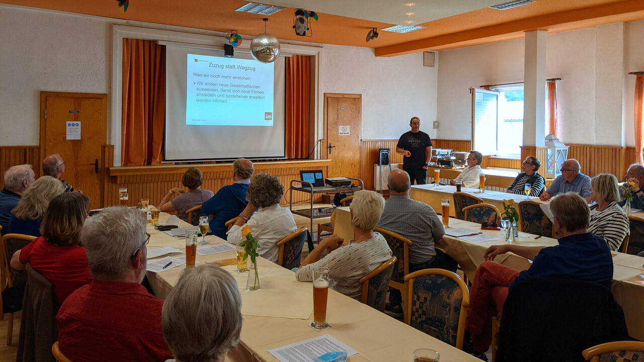 Menschen sitzen im Saal des Lokals Goldene Aue vor einer Leinwand, auf der eine Präsentation projeziert wird. 